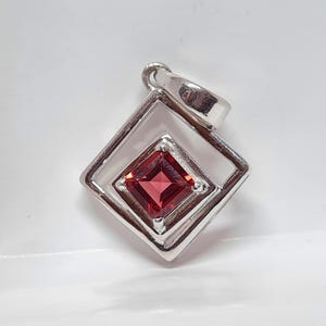 Op de afbeelding: Zilveren hanger met een vierkante, dieprode edelsteen, gezet in een ruitvormige omlijsting. De hanger heeft een eenvoudig, elegant ontwerp. De edelsteen heeft een levendige, rijke rode kleur.