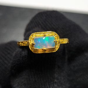 May include: A gold ring featuring a rectangular opal gemstone with iridescent colors of blue, green, and pink. The ring has an ornate, filigree-style setting and a delicate band.