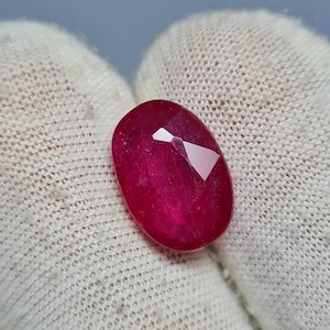 May include: A faceted, oval-shaped ruby gemstone. The gem is a deep, rich red color with multiple facets that catch the light. The ruby is held against a white, textured background.