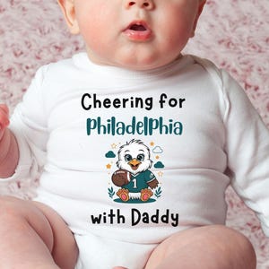 K&ouml;nnte beinhalten: Ein wei&szlig;er, lang&auml;rmeliger Babybody mit dem Text "Cheering for Philadelphia with Daddy" in Schwarz und T&uuml;rkis. Unter dem Text befindet sich eine Grafik eines Cartoon-Adlers, der ein t&uuml;rkisfarbenes Football-Trikot mit der Nummer 1 tr&auml;gt.