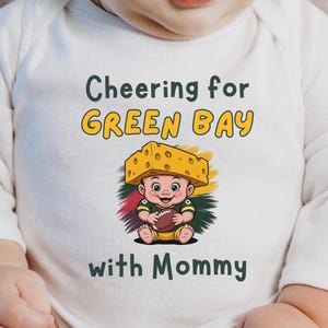 May include: White baby bodysuit with long sleeves. The front features the text "Cheering for GREEN BAY with Mommy" above a cartoon baby wearing a cheesehead and holding a football. The design includes green, yellow, red, and brown colors.
