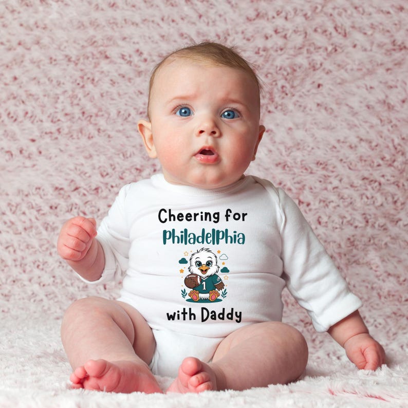 K&ouml;nnte beinhalten: Ein wei&szlig;er, lang&auml;rmeliger Babybody mit dem Text "Cheering for Philadelphia with Daddy" in Schwarz und T&uuml;rkis. Unter dem Text befindet sich eine Grafik eines Cartoon-Adlers, der ein t&uuml;rkisfarbenes Football-Trikot mit der Nummer 1 tr&auml;gt.