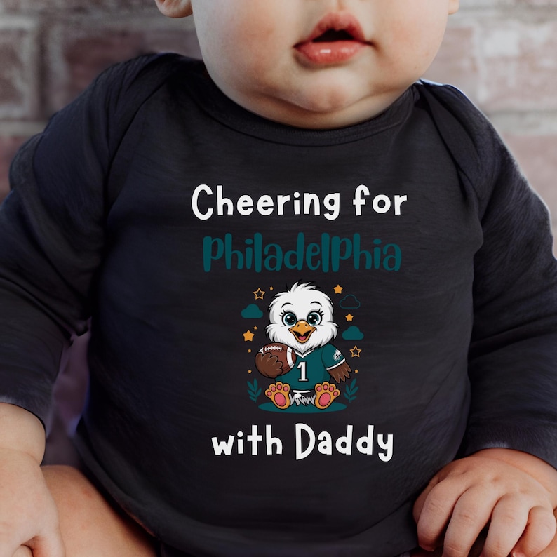 K&ouml;nnte beinhalten: Ein schwarzer lang&auml;rmeliger Babybody mit dem wei&szlig;en Text Cheering for, dem blaugr&uuml;nen Text Philadelphia und dem wei&szlig;en Text with Daddy. Eine Grafik eines Cartoon-Adlers, der ein Trikot mit der Nummer 1 tr&auml;gt und einen Football h&auml;lt.