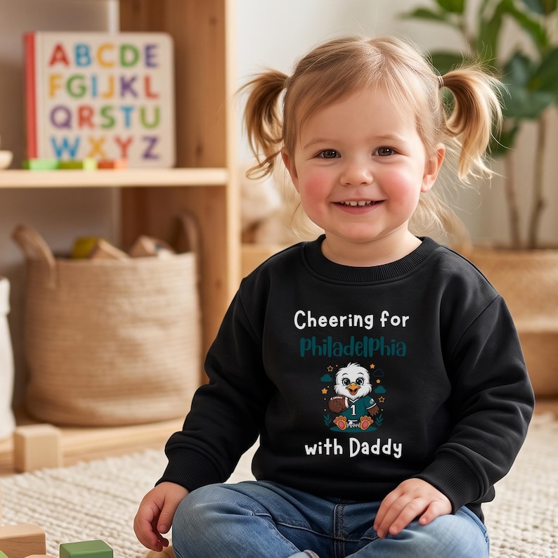 K&ouml;nnte beinhalten: Ein schwarzes Langarm-Sweatshirt mit einer Grafik eines Cartoon-Adlers, der ein Trikot mit der Nummer 1 tr&auml;gt und einen Football h&auml;lt. Der Text Cheering for Philadelphia with Daddy ist in wei&szlig;er und t&uuml;rkiser Schrift aufgedruckt.