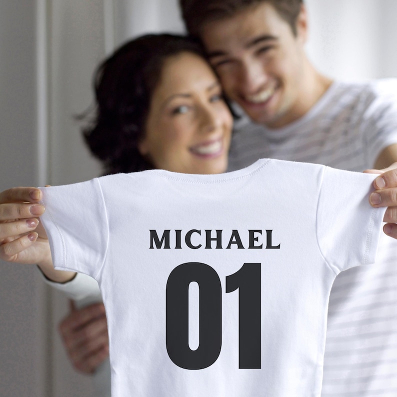 K&ouml;nnte beinhalten: Ein wei&szlig;er kurz&auml;rmeliger Babybody mit dem Namen MICHAEL in schwarzen Gro&szlig;buchstaben &uuml;ber der gro&szlig;en schwarzen Zahl 01. Das Kleidungsst&uuml;ck besteht aus schlichtem wei&szlig;em Stoff und zeigt den Text deutlich im vorderen Brustbereich.