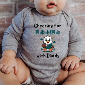 K&ouml;nnte beinhalten: Ein lang&auml;rmeliger Babybody in Grau meliert mit dem Text "Cheering for Philadelphia with Daddy" in Schwarz und Petrol. Die Grafik in der Mitte zeigt einen Cartoon-Adler, der ein petrolfarbenes Trikot mit der Nummer 1 tr&auml;gt.