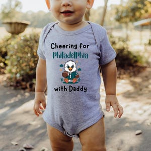 K&ouml;nnte beinhalten: Ein graumelierter kurz&auml;rmeliger Babybody mit dem schwarzen Text Cheering for Philadelphia with Daddy und einer Grafik eines Cartoon-Adlers, der ein Trikot mit der Nummer 1 tr&auml;gt und einen Football h&auml;lt, umgeben von kleinen Wolken.