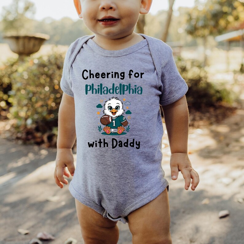 K&ouml;nnte beinhalten: Ein graumelierter kurz&auml;rmeliger Babybody mit dem schwarzen Text Cheering for Philadelphia with Daddy und einer Grafik eines Cartoon-Adlers, der ein Trikot mit der Nummer 1 tr&auml;gt und einen Football h&auml;lt, umgeben von kleinen Wolken.