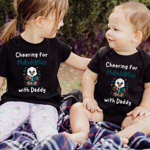 K&ouml;nnte beinhalten: Ein schwarzes kurz&auml;rmeliges T-Shirt und ein schwarzer kurz&auml;rmeliger Body, beide mit dem wei&szlig;en und blaugr&uuml;nen Schriftzug "Cheering for Philadelphia with Daddy" und einer Grafik eines Adlers im Football-Trikot mit Football.