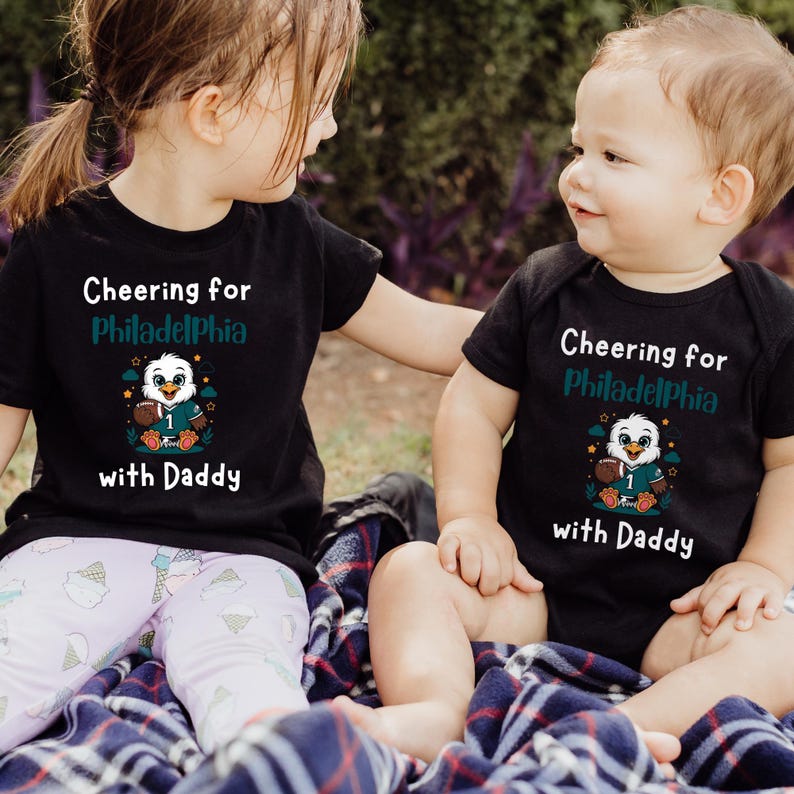 K&ouml;nnte beinhalten: Ein schwarzes kurz&auml;rmeliges T-Shirt und ein schwarzer kurz&auml;rmeliger Body, beide mit dem wei&szlig;en und blaugr&uuml;nen Schriftzug "Cheering for Philadelphia with Daddy" und einer Grafik eines Adlers im Football-Trikot mit Football.