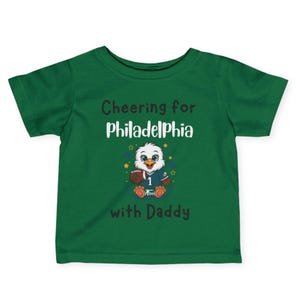 K&ouml;nnte beinhalten: Ein dunkelgr&uuml;nes Kurzarm-T-Shirt mit einem Cartoon-Adler, der ein Trikot mit der Nummer 1 tr&auml;gt und einen Football h&auml;lt. Der Text lautet Cheering for Philadelphia with Daddy in schwarzer und wei&szlig;er Schrift, umgeben von kleinen Sternen.