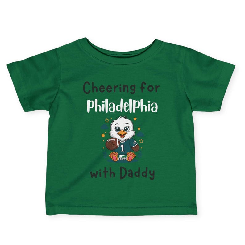 K&ouml;nnte beinhalten: Ein dunkelgr&uuml;nes Kurzarm-T-Shirt mit einem Cartoon-Adler, der ein Trikot mit der Nummer 1 tr&auml;gt und einen Football h&auml;lt. Der Text lautet Cheering for Philadelphia with Daddy in schwarzer und wei&szlig;er Schrift, umgeben von kleinen Sternen.