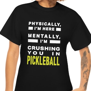 Puede incluir: Camiseta negra con texto blanco que dice "Físicamente, estoy aquí. Mentalmente, te estoy aplastando en pickleball." El texto está en un cuadro con borde amarillo.