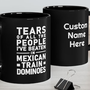 May include: Two black ceramic mugs. One mug has the text "TEARS OF ALL THE PEOPLE I'VE BEATEN IN MEXICAN TRAIN DOMINOES" in white. The other mug has the text "Custom Name Here" in white.