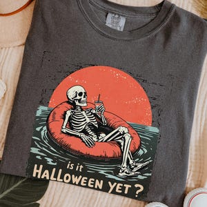 Könnte beinhalten: Dunkelgraues T-Shirt mit einem Skelett, das sich auf einem roten Schwimmring im Wasser entspannt, mit einer roten Sonne im Hintergrund. Der Text "Is it Halloween Yet?" ist darunter. Ein lässiges T-Shirt für Halloween.
