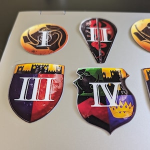 Puede incluir: Cinco pegatinas de escudo de color con los números romanos I, II, III, IV y una corona. Las pegatinas están en un portátil plateado.
