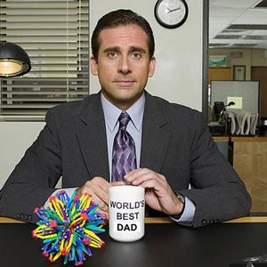 May include: A white ceramic mug with the text "World's Best Dad" in black lettering. The mug is being held by a person wearing a dark suit and a patterned tie. A colorful, multi-faceted, sphere-shaped toy is in the foreground.
