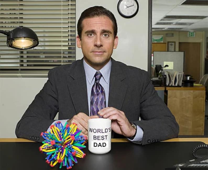 May include: A white ceramic mug with the text "World's Best Dad" in black lettering. The mug is being held by a person wearing a dark suit and a patterned tie. A colorful, multi-faceted, sphere-shaped toy is in the foreground.