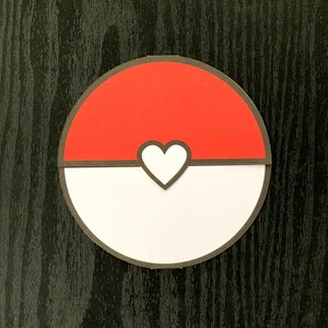 May include: A circular flat object featuring a red top half and a white bottom half, separated by a horizontal black line. A white heart shape with a black outline is centred on the horizontal line, resembling a stylised ball design.