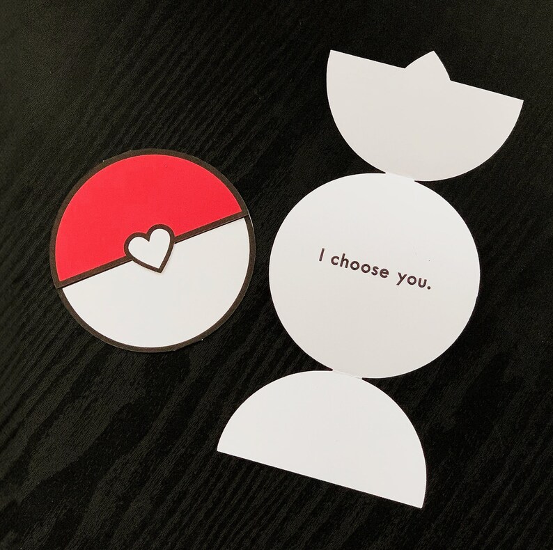 May include: A circular card featuring a red and white design with a central heart, resembling a ball. Beside it, an unfolded white card displays the text I choose you in dark brown lettering, with semi-circular flaps at the top and bottom.