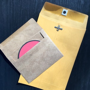 May include: A large yellow envelope with a metal clasp closure and a smaller brown kraft paper envelope containing a circular pink card with a black border. The yellow envelope features a reinforced white eyelet for the metal fastener.