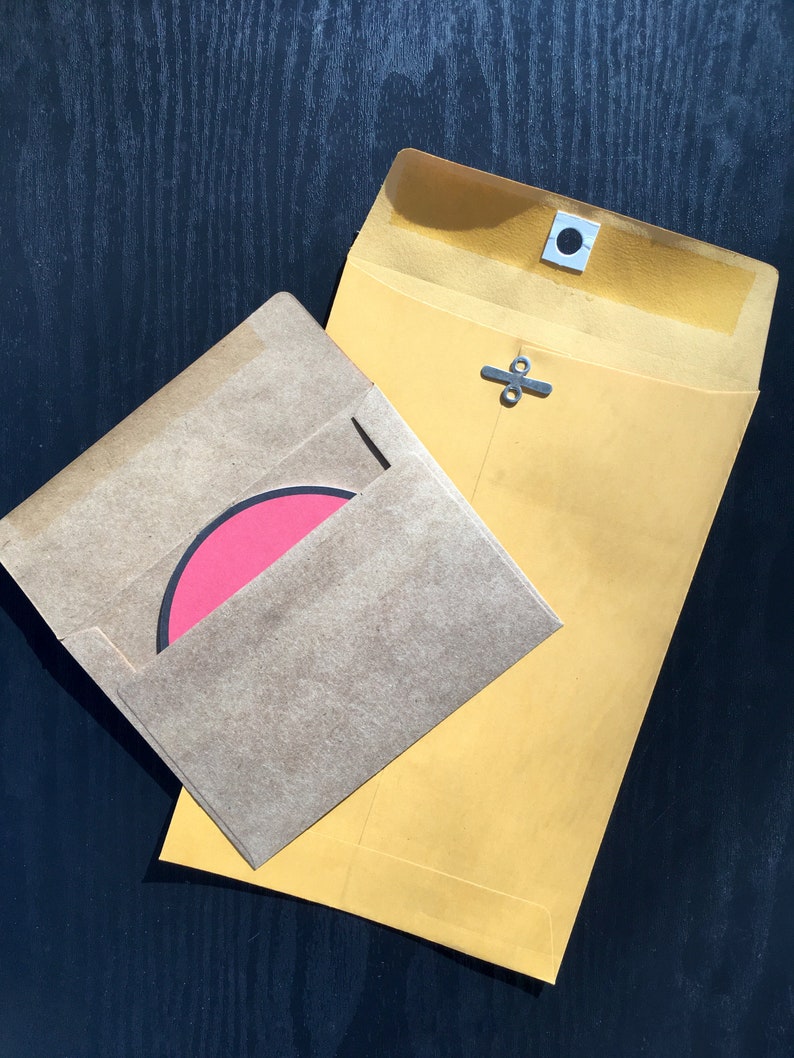 May include: A large yellow envelope with a metal clasp closure and a smaller brown kraft paper envelope containing a circular pink card with a black border. The yellow envelope features a reinforced white eyelet for the metal fastener.