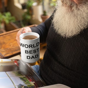 May include: White ceramic mug with black text that reads "World Best Dad". A person is holding the mug and reading a book.