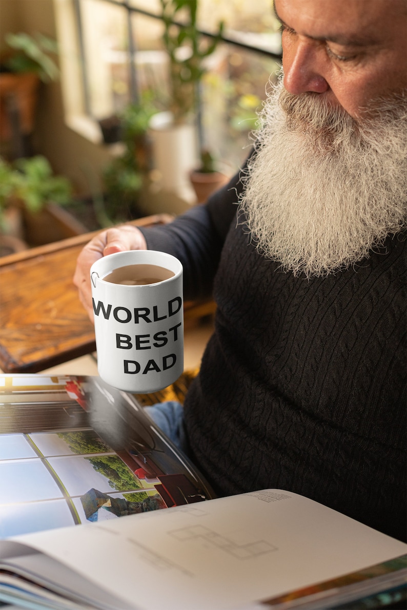 May include: White ceramic mug with black text that reads "World Best Dad". A person is holding the mug and reading a book.