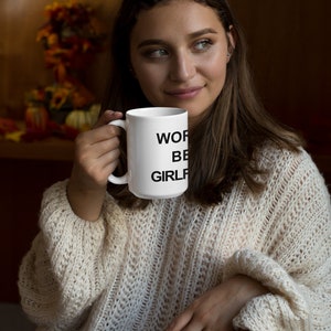 May include: White ceramic mug with black text that reads "Work, Be, Girlf". A person is holding the mug in their right hand.