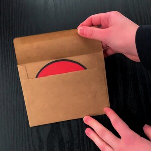 May include: A square brown paper envelope with an open flap, revealing a circular object with a bright red centre and a thin black border partially inserted inside the envelope, held against a dark wood-grain surface.
