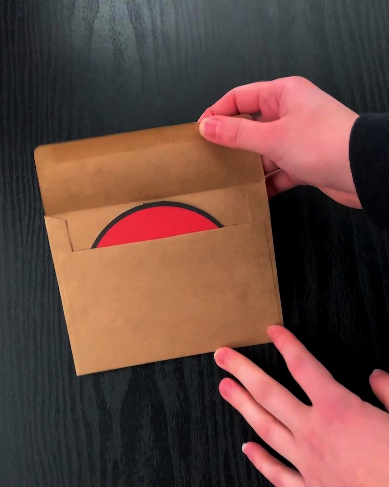 May include: A square brown paper envelope with an open flap, revealing a circular object with a bright red centre and a thin black border partially inserted inside the envelope, held against a dark wood-grain surface.