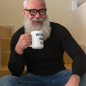 May include: A man with a white beard and glasses is sitting on stairs holding a white mug with the text "Work, Be, Done" printed on it.