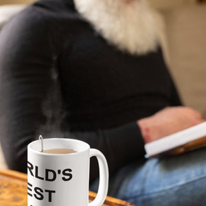 May include: White ceramic mug with black text that reads "WORLD'S BEST DAD". The mug is filled with a brown liquid and has steam rising from it. The mug is sitting on a wooden surface.