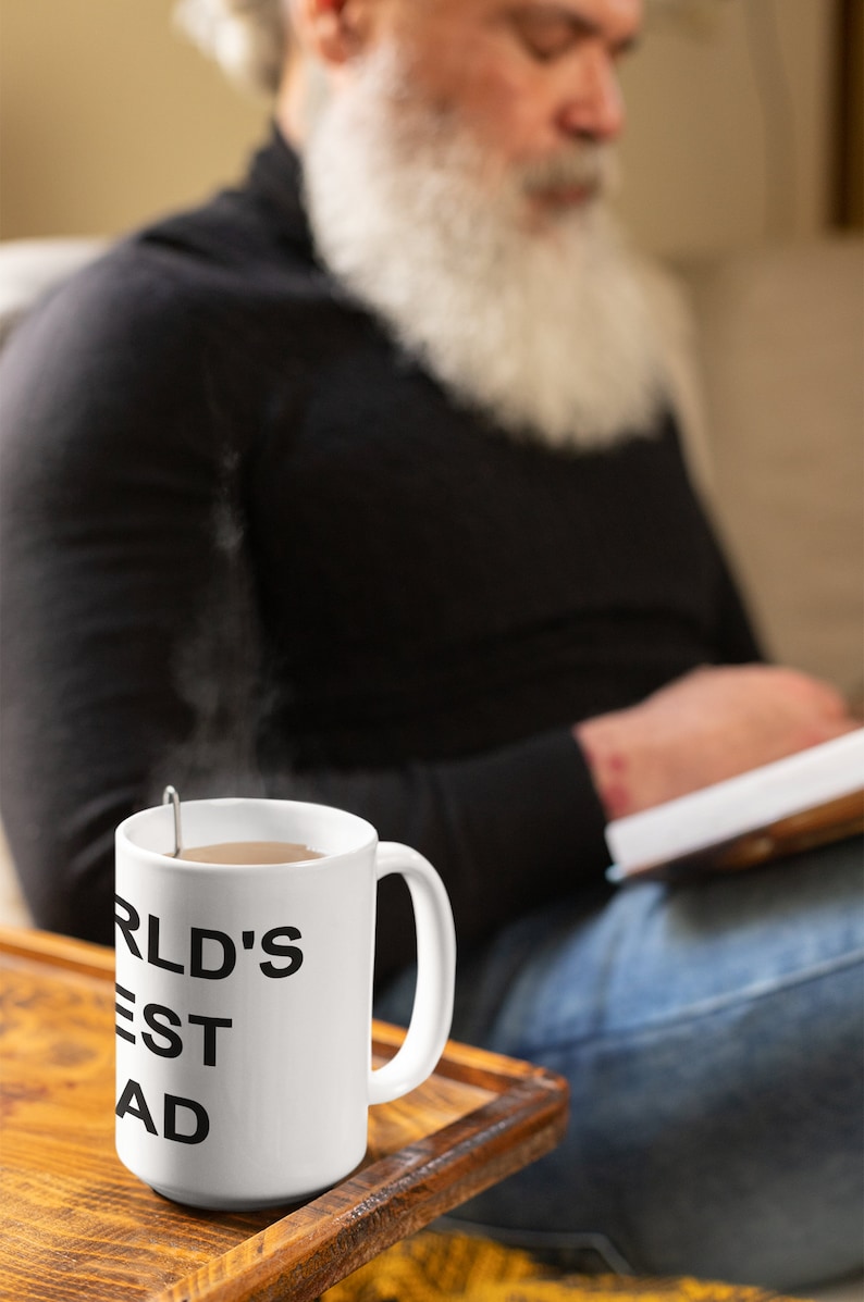 May include: White ceramic mug with black text that reads "WORLD'S BEST DAD". The mug is filled with a brown liquid and has steam rising from it. The mug is sitting on a wooden surface.