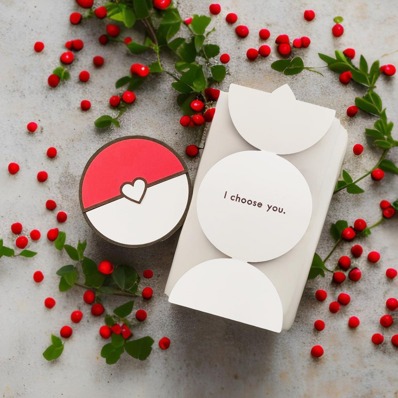May include: A circular red and white container with a heart-shaped centre detail next to a rectangular white box featuring a circular flap printed with the text I choose you. The items are surrounded by small red berries and green leaves.