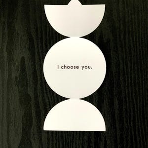 May include: A white paper card with a vertical design consisting of a top semi-circle, a central full circle, and a bottom semi-circle. The central circle features the black printed text "I choose you." in a simple, sans-serif font.