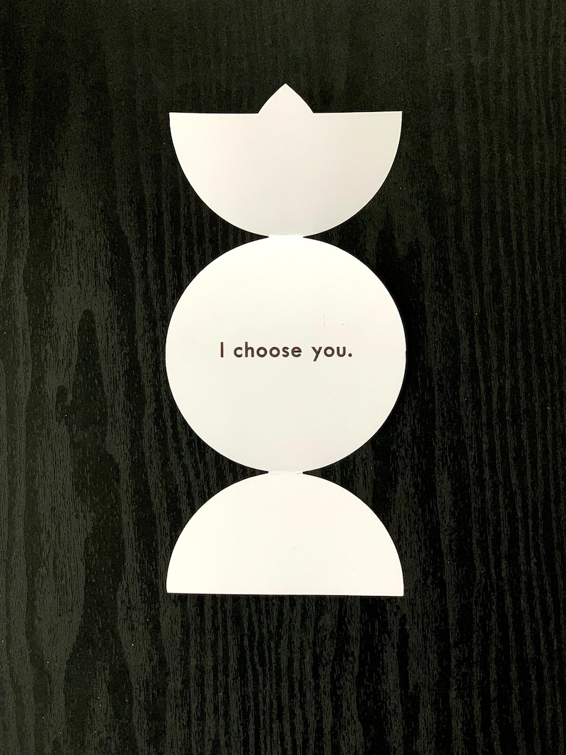May include: A white paper card with a vertical design consisting of a top semi-circle, a central full circle, and a bottom semi-circle. The central circle features the black printed text "I choose you." in a simple, sans-serif font.