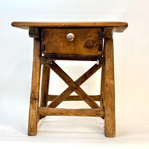 May include: A rustic wooden side table with a single drawer. The table features a natural wood finish, a rectangular top, and a drawer with a white knob. The legs are joined by an X-shaped support, creating a sturdy base. The table measures approximately 24 inches high.