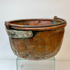 May include: A weathered copper bucket with a rounded body and a sturdy handle. The bucket has a tarnished patina with hints of green and a riveted metal band. The rim is reinforced with a metal band.