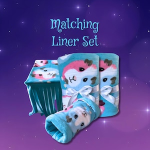 May include: A matching liner set featuring a light blue fleece fabric with a repeating pattern of pink and white guinea pig faces. The set includes a fringed rectangular mat, two rolled pieces tied with white ribbons, and a small tunnel-shaped piece.