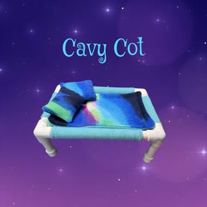 May include: A Cavy Cot pet bed featuring a white PVC pipe frame with light blue fleece covers. The bed includes a rectangular fleece mat and a small pillow, both decorated with a vibrant blue, green, and purple nebula-inspired galaxy print.