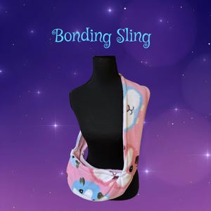 May include: A pink fleece bonding sling featuring a repeating pattern of blue and white cartoon animals. The text Bonding Sling is printed in light blue script above the sling, which is displayed on a black mannequin torso.