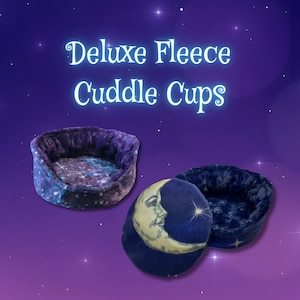 May include: Two round fleece cuddle cups for small pets. One cup features a purple galaxy print with a textured interior. The other cup is dark blue with a crescent moon and star graphic on the base. The text Deluxe Fleece Cuddle Cups is visible.