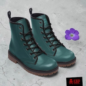 May include: A pair of teal combat boots with black laces and a skull emblem. The boots have a brown sole and a pull tab at the back. A purple flower rests nearby. The boots are a stylish and edgy footwear option.
