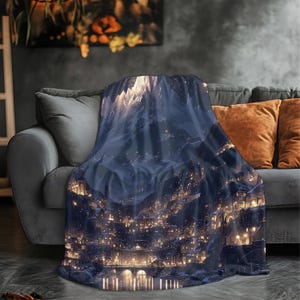 May include: A soft throw blanket with a digital art print depicting a mountain village at night. The design shows a detailed landscape with illuminated buildings, a bridge, and a reflective body of water, in shades of blue and gold.