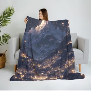 May include: A dark blue blanket with a detailed illustration of a mountain range at night. The artwork depicts a city in the valley below, with many illuminated buildings and a starry sky. The blanket is draped over a gray sofa.