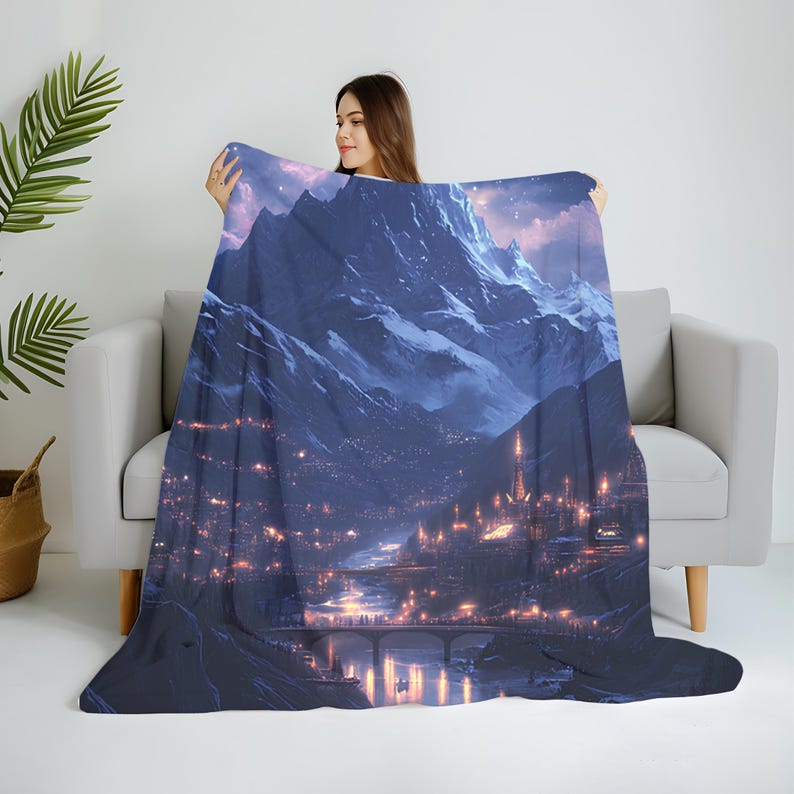 May include: A plush blanket featuring a detailed illustration of a mountain range at dusk. The artwork depicts a city with illuminated buildings, a bridge, and a river, all under a starry sky. The color palette includes shades of blue, purple, and orange.