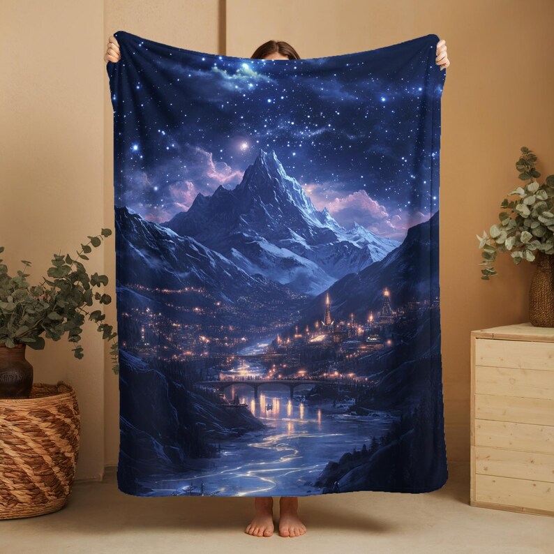 May include: A dark blue blanket featuring a detailed illustration of a mountain range under a starry night sky. The artwork depicts a river, a bridge, and a city with illuminated buildings. The blanket is held up, showcasing the full design.