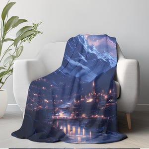May include: A plush throw blanket draped over a white armchair. The blanket features a digital art print of a nighttime cityscape nestled at the foot of snow-capped mountains. The city lights glow warmly against the cool blue tones.