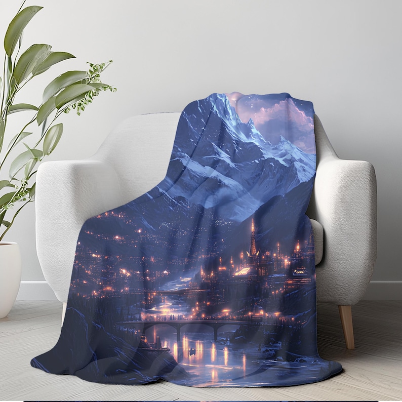 May include: A plush throw blanket draped over a white armchair. The blanket features a digital art print of a nighttime cityscape nestled at the foot of snow-capped mountains. The city lights glow warmly against the cool blue tones.